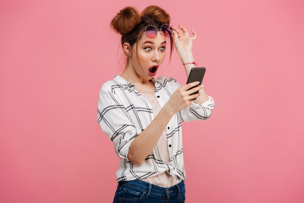 Portrait of a shocked young girl using mobile phone isolated over pink background